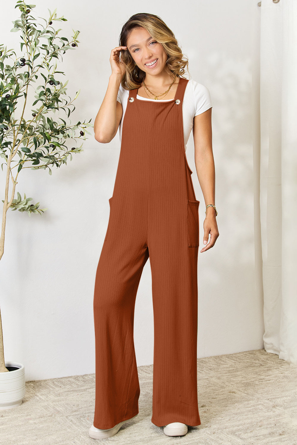 Woman wearing brown wide strap overall with pockets and a white shirt, standing in a stylish indoor setting.