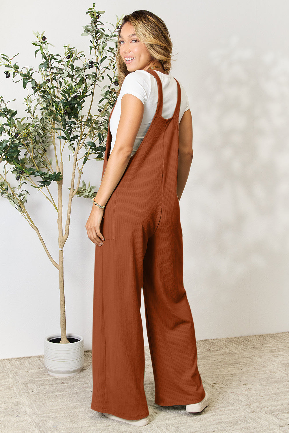 Woman wearing wide strap overall with pockets in terracotta color, standing next to a potted plant.