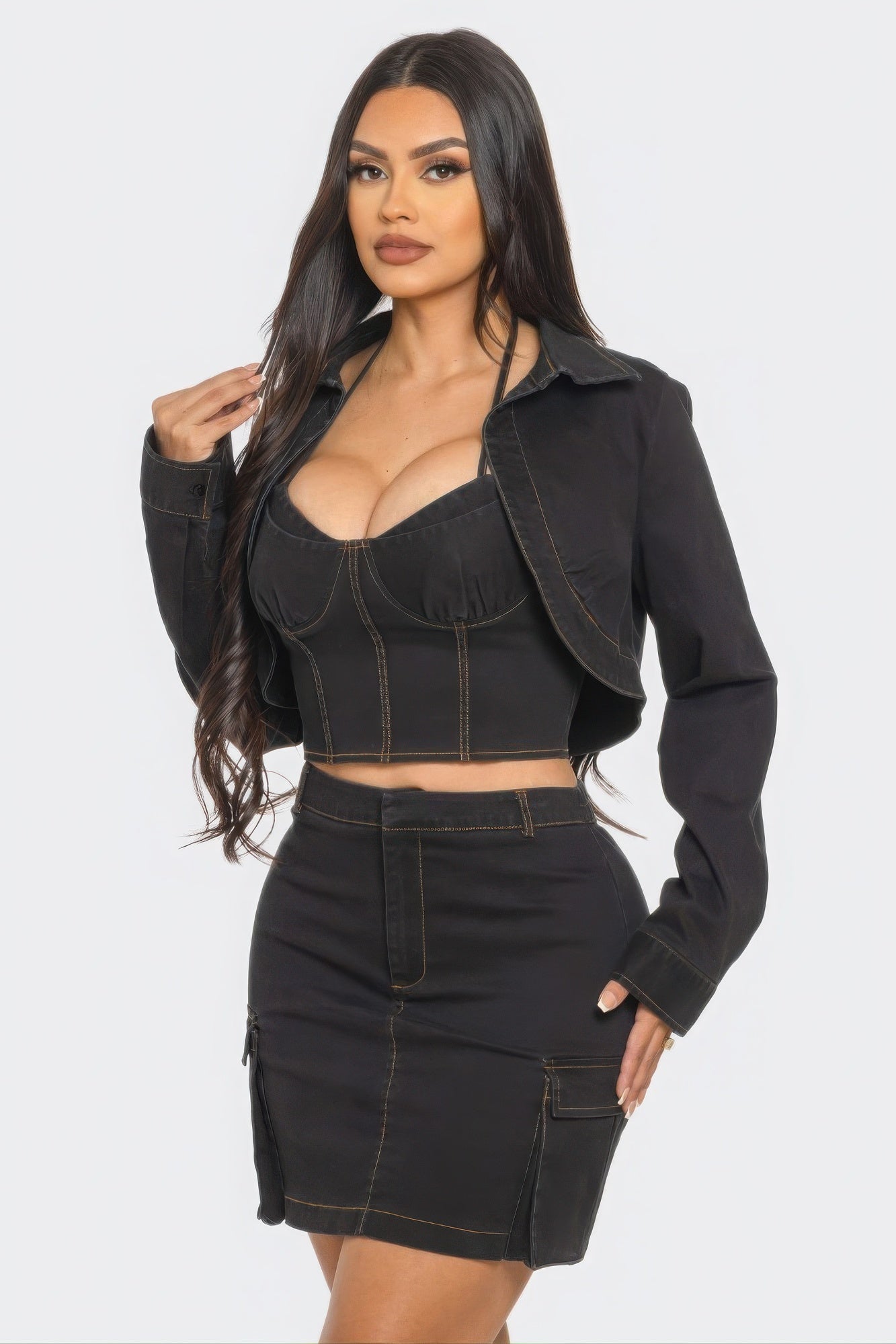 Woman with long dark hair wearing a black denim three-piece outfit consisting of a cropped bustier top, matching jacket, and mini skirt with contra...