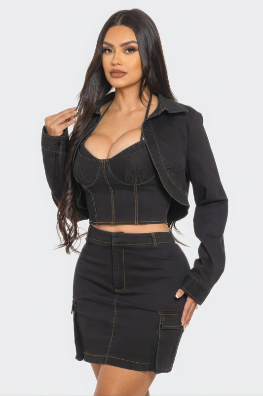 Woman with long dark hair wearing a black denim three-piece outfit consisting of a cropped bustier top, matching jacket, and mini skirt with contra...