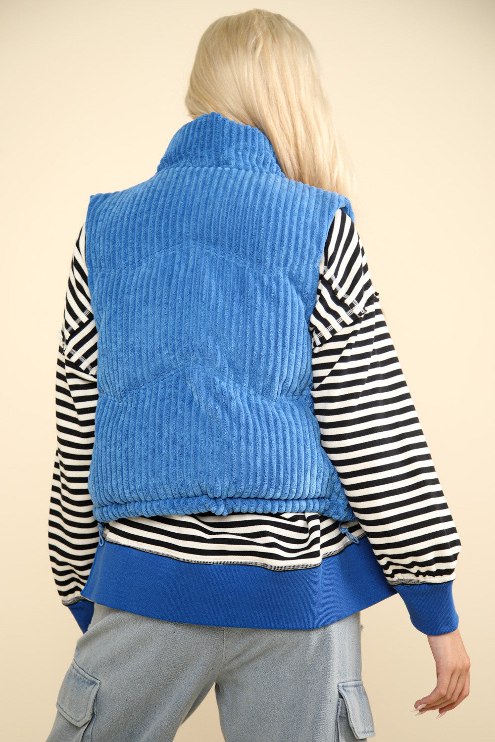 Woman wearing blue corduroy puffer vest with zig-zag pattern, shown from back, layered over striped sweater and jeans.