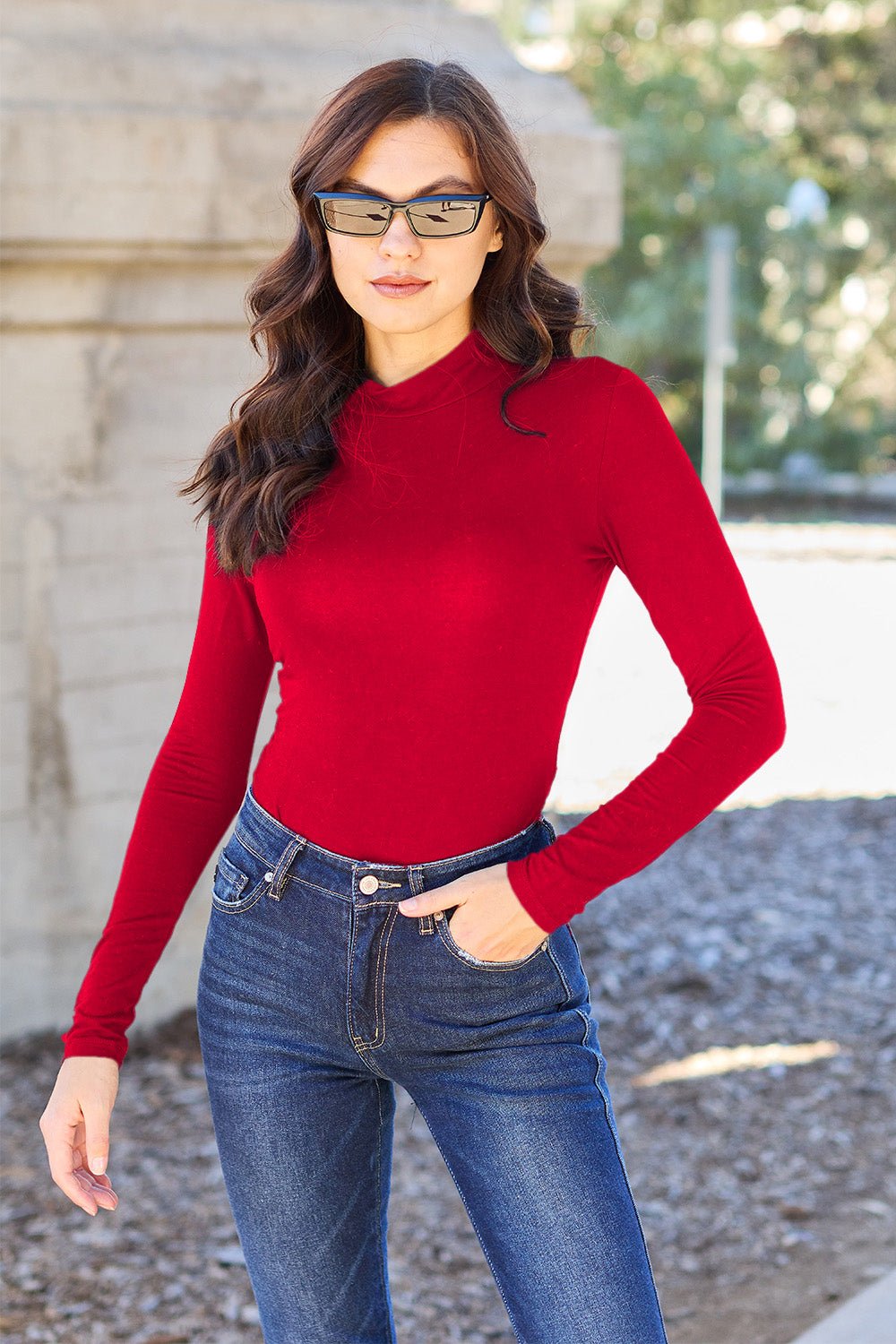 Woman wearing red mock neck long sleeve bodysuit with jeans, sunglasses, standing outdoors in casual style.