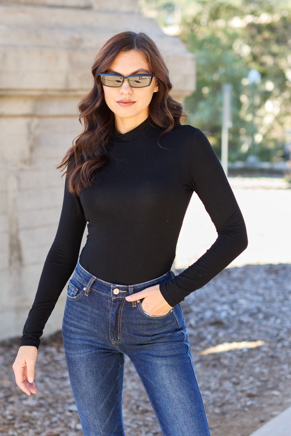Woman in black mock neck long sleeve bodysuit and jeans standing outdoors wearing sunglasses.