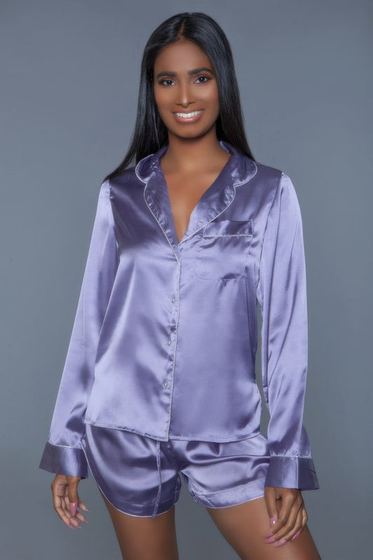 Woman wearing purple satin pajama set with button-up shirt and shorts, smiling at camera against gray background.