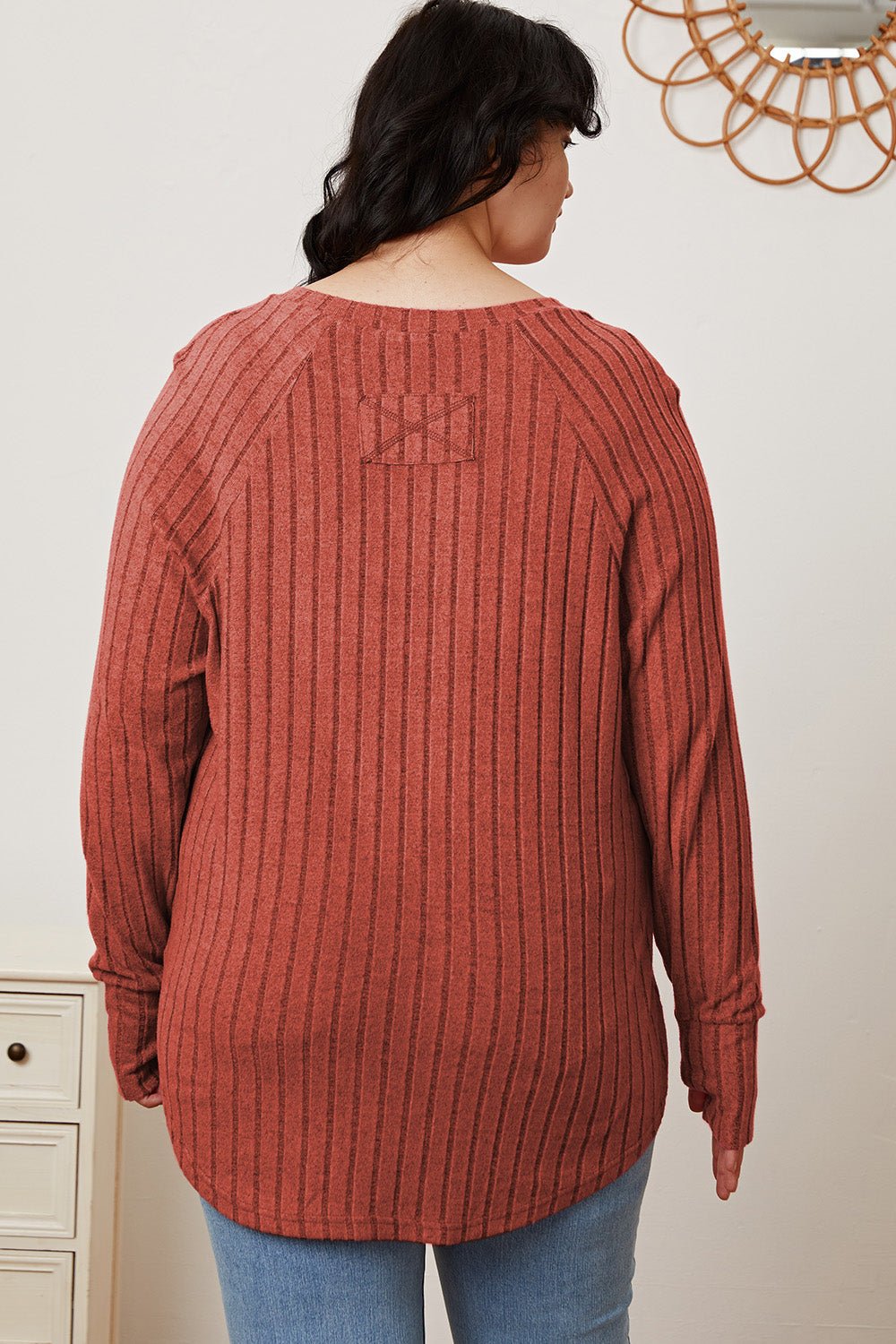 Woman wearing a ribbed, rust-colored long sleeve t-shirt with thumbholes, showing back view standing in a casual room setting.