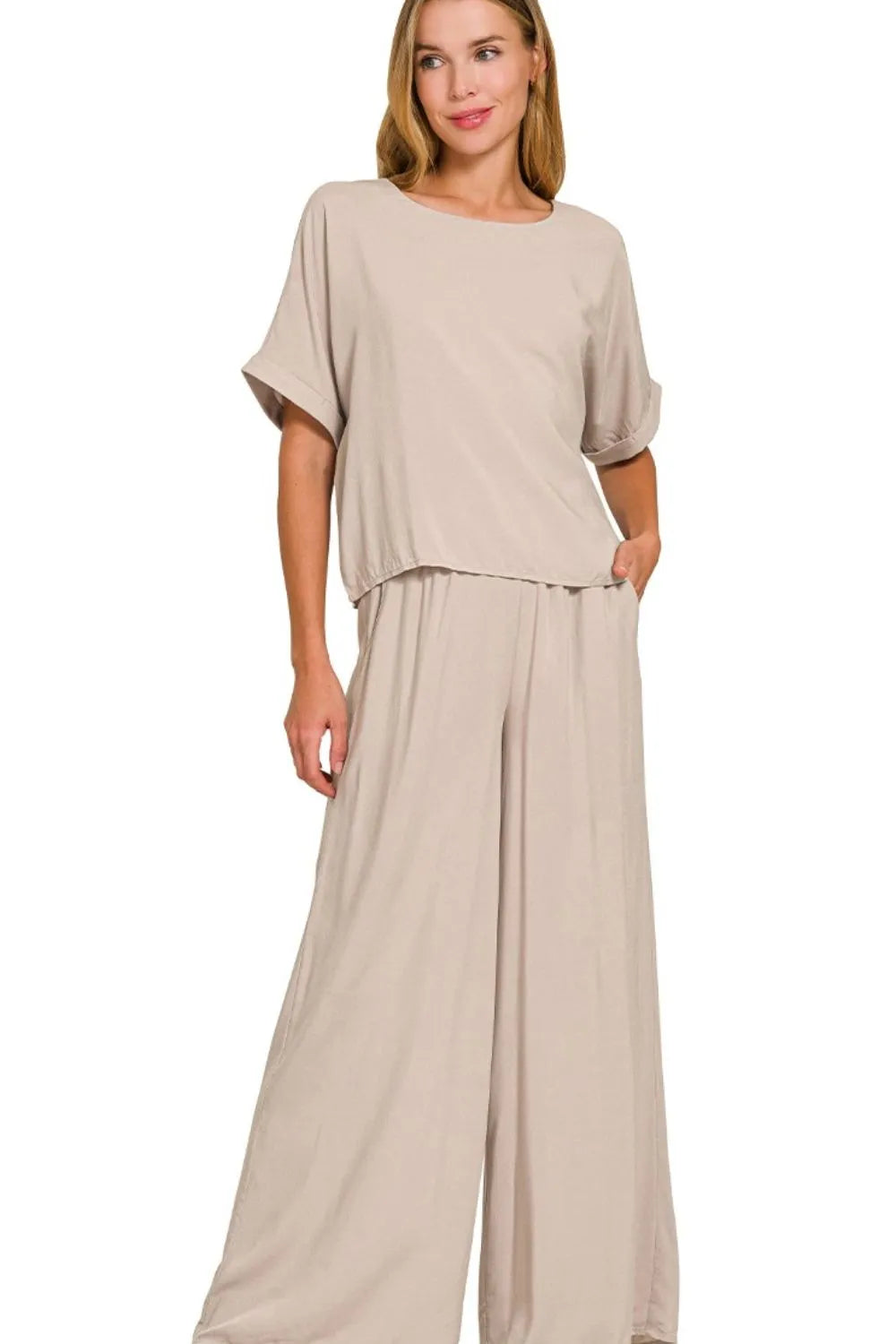 Woman wearing a beige two-piece loungewear set with oversized short-sleeve top and wide-leg pants, standing against white background with hand in p...