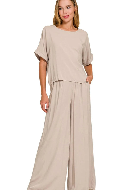 Woman wearing a beige two-piece loungewear set with oversized short-sleeve top and wide-leg pants, standing against white background with hand in p...